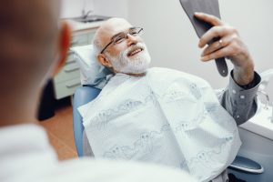 A man with a white beard smiles in a handheld mirror at the results of a cosmetic dentistry procedure. He's in a dentist chair.